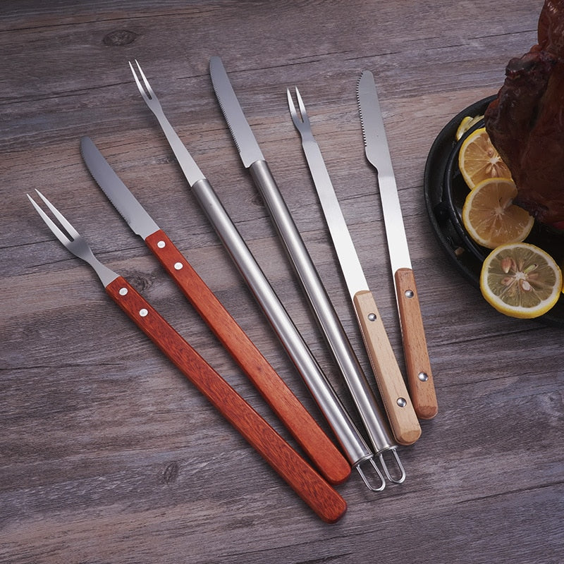 Stainless steel roast lamb knife and fork set on a wooden table with barbecue tool.